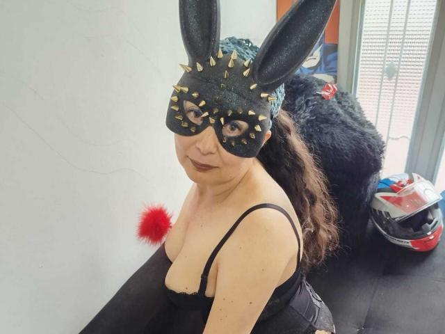 BrianDance with black mask, spiked ears, and black dress, showing tits, sitting on black couch, black furry dog behind, helmet with red pom-pom, and window in background.
