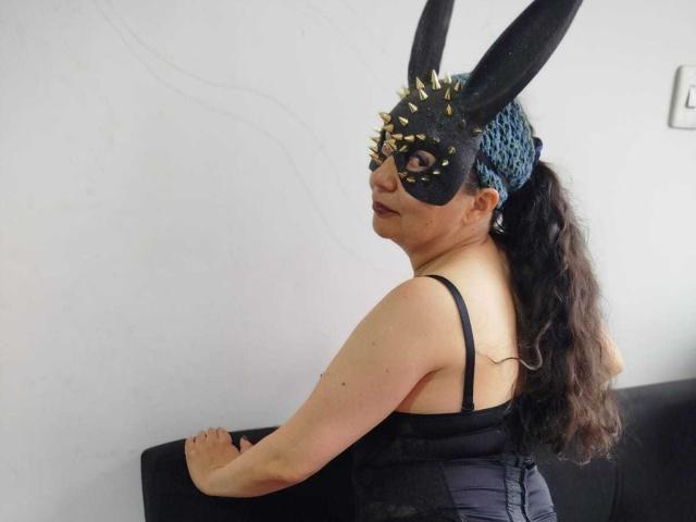 BrianDance with long brown hair in a ponytail, wearing a black mask with spikes, black corset, and black bunny ears, sitting on a black couch, looking back.