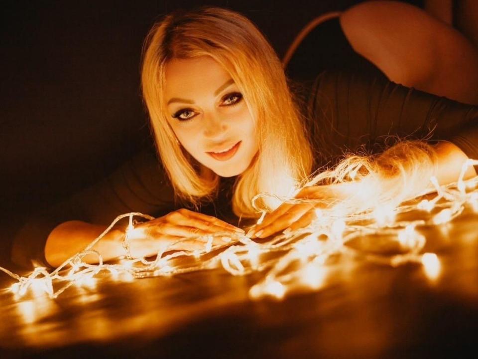 XPrettyQueenX lies on a dark surface, surrounded by glowing fairy lights. Her blonde hair is straight and falls over her shoulders. She has a warm, inviting smile and wears a dark green long-sleeve top. The light from the fairy lights illuminates her face and hands, creating a soft, golden glow. The focus is on her face and the lights, with her hands resting on the lights. The composition is warm and intimate, with a strong contrast between the dark background and the bright fairy lights.