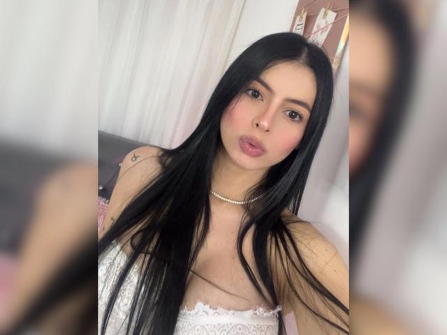 IsabellaJames met lang zwart haar, volle roze lippen, een witte kanten top, kleine borsten, een slank figuur, een ketting, een tatoeage op de linker schouder, een onscherpe achtergrond, een spiegelzelfportret..