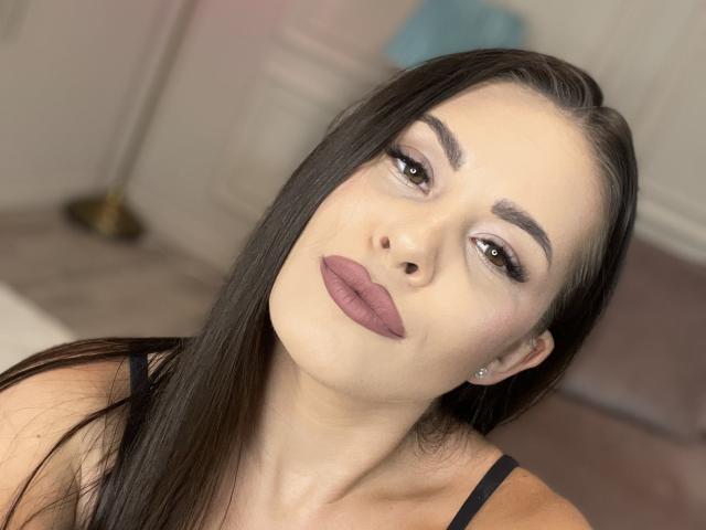 KatieHayes has long, straight black hair and brown eyes. She wears bold, matte pink lipstick and subtle, smoky eye makeup. Her eyebrows are thick and perfectly shaped. She has a small silver stud in her left ear. KatieHayes's skin is fair, and she looks directly at the camera with a slightly tilted head. She wears a black strap, possibly from a top, is visible on her shoulder. The background is blurred, featuring a soft, beige carpet and a blue, fuzzy object in the back. The lighting is even, highlighting her facial features.