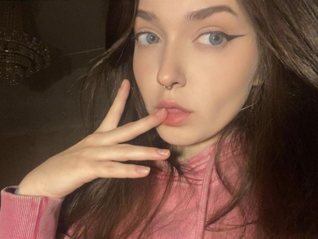 IrishReversed with pouty lips, nose ring, and winged eyeliner, touching her finger to her pink lips, wearing a pink turtleneck, has long brown hair and blue eyes.