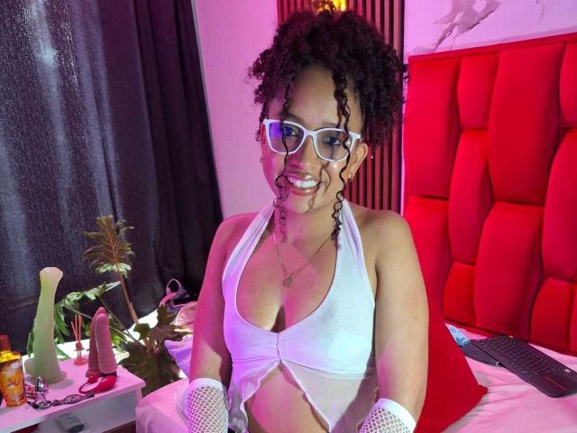 ArtemisaS with curly black hair, white glasses, and white cut-out top showing her tits. She's smiling with pink and purple lighting, wearing white fishnet gloves, in a bedroom.