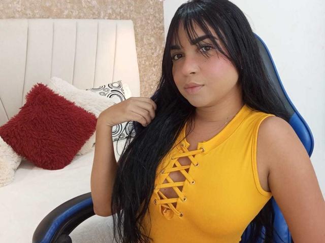 AliceSmity with long black hair, yellow lace-up top, small tits, sitting in blue chair, hand in hair, white wall, beige patterned background, red and white pillows.
