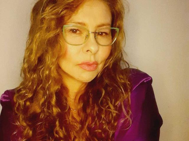 MiaAmoretti with wavy brown hair, green glasses, and pouty lips, wearing a purple velvet top, has a slight facial blemish, and a small necklace.