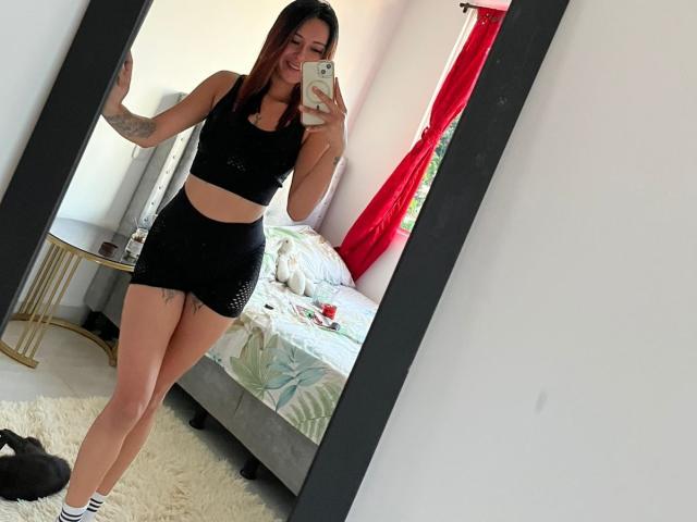 DamiAmbani with brown hair, smiling, taking a mirror selfie. She's wearing a black sports bra and shorts, showing her tits and ass, with tattoos on her arm.