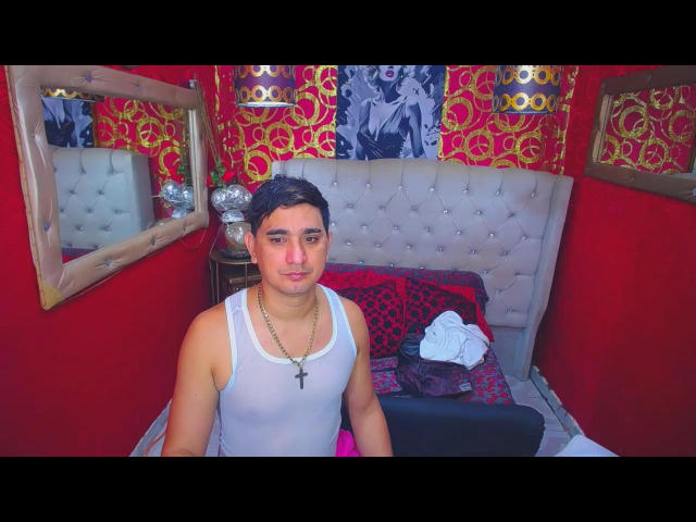 DuoedJhova, chubby, short black hair, wearing white tank top, black cross necklace, sitting on bed, red walls, tufted gray headboard, red and black pillows, white cloth on bed.
