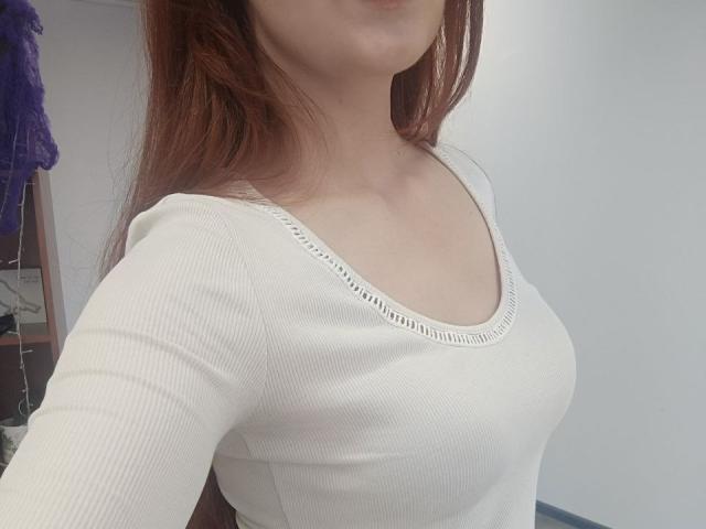 Close-up of Gvineriya's upper body. She has light skin and long, straight, reddish-brown hair. She wears a white, ribbed, long-sleeve top with a slightly scalloped edge on the neckline. Her small to medium-sized breasts are clearly visible through the tight fabric. The image is a photograph with a simple, neutral background. The texture of the top is smooth and slightly shiny. The composition focuses on her chest and face, with her face partially out of the frame.