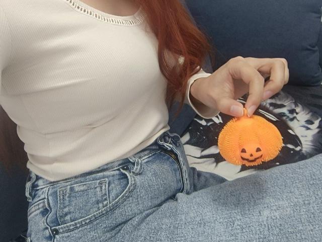 Gvineriya with red hair, white ribbed shirt, and blue jeans, holding an orange fluffy pumpkin toy with a cute face, on a dark blue couch.