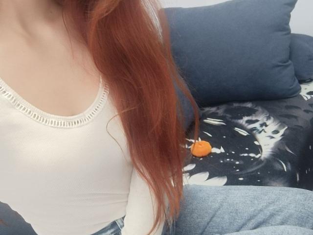 Gvineriya mit roten Haaren, die ein weißes, geripptes Tanktop trägt und ihre kleinen Brüste zeigt, sitzt in blauen Jeans auf einem schwarz-weißen Sofa mit einem orangefarbenen Gegenstand..