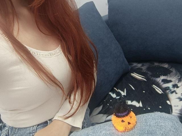 Gvineriya mit roten Haaren, einem weißen, gerippten Oberteil und blauen Jeans, sitzt auf einem dunkelblauen Sofa, mit einem kleinen, flauschigen, orangefarbenen Kürbis-Spielzeug im Schoß..