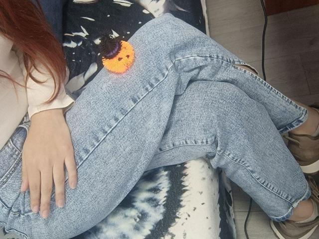 Gvineriya's lower half is in the image. She wears loose, light blue denim pants with a small, orange and purple floral patch on the right thigh. Her right hand rests on her pants. She has long red hair, and her white sleeve covers her left arm. Brown sandals are visible. The background includes a tie-dye patterned blanket and a wooden floor. The image has a casual, relaxed feel.