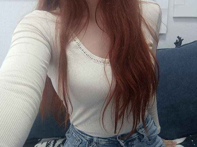Gvineriya mit langen, roten Haaren, die ein weißes, geripptes Langarmshirt trägt und ihre Brüste in hoch taillierten, blauen Jeans zeigt, sitzt auf einem dunkelblauen Sofa..