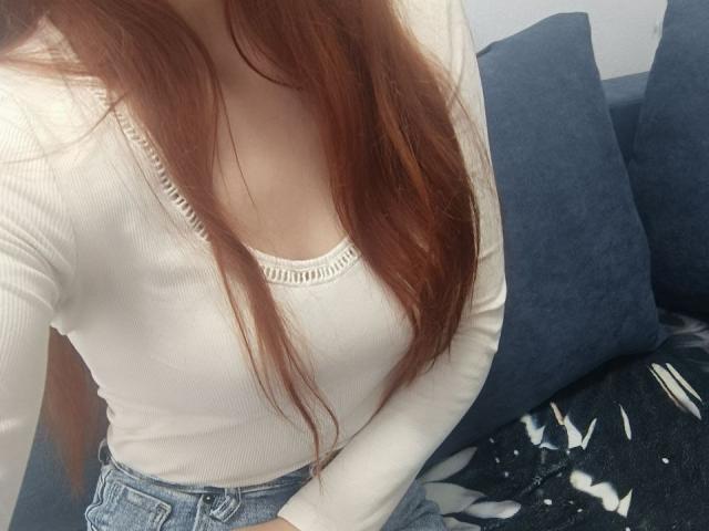 Gvineriya mit langen, roten Haaren, einem weißen, gerippten Langarmshirt, das kleine Brüste zeigt, und blauen Jeans, sitzt auf einem dunkelblauen Sofa mit einer schwarz-weißen Decke..