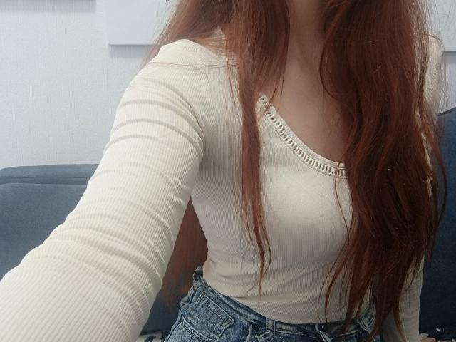 Gvineriya mit langen, roten Haaren, die ein weißes, geripptes Langarmshirt und blaue Jeans trägt, wobei kleine Brüste und eine schlanke Taille zu sehen sind. Sie macht ein Selfie..
