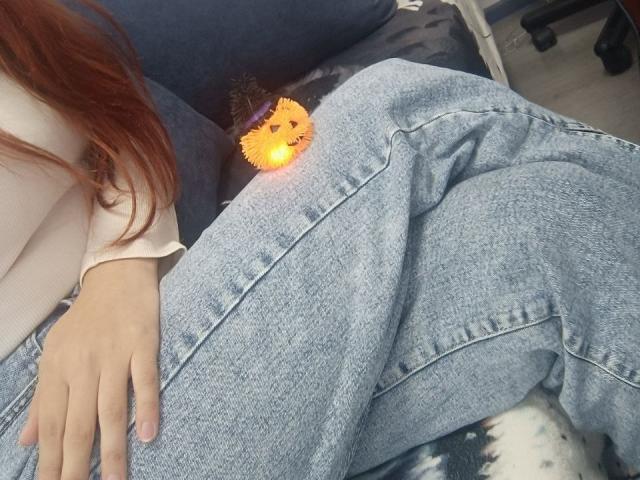 A close-up photo of Gvineriya's lower body. She has long red hair and wears light blue, slightly baggy denim pants. Her right hand rests on her thigh. A small, glowing, orange emoji with a happy face is positioned near her hip. The texture of the denim is rough and visible. The background includes a black and white patterned blanket. Gvineriya's skin is light. The image uses natural lighting.