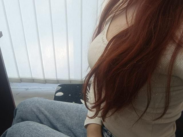 Gvineriya mit langen, roten Haaren, einem weißen, gerippten Oberteil, das ihre Brüste zeigt, sitzt in blauen Jeans, in der Nähe eines schwarzen, gepunkteten Kissens, vor einem Fenster mit vertikalen Jalousien..