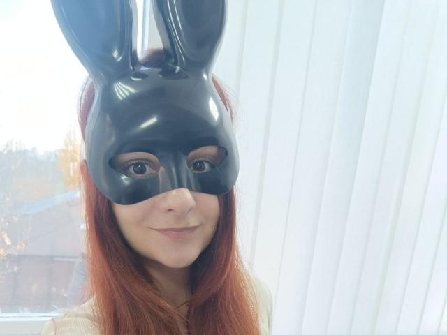 Gvineriya mit roten Haaren, die eine glänzende schwarze Hasenmaske trägt und leicht lächelt, im Hintergrund weiße, vertikale Jalousien, ein Fenster mit unscharfem Außenlicht..