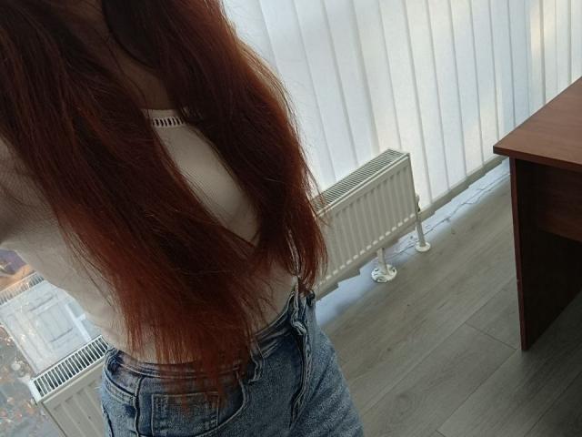 Gvineriya has long red hair, a low-cut white top, and blue jeans, showing her tits and a bit of her ass. She's standing next to a radiator.