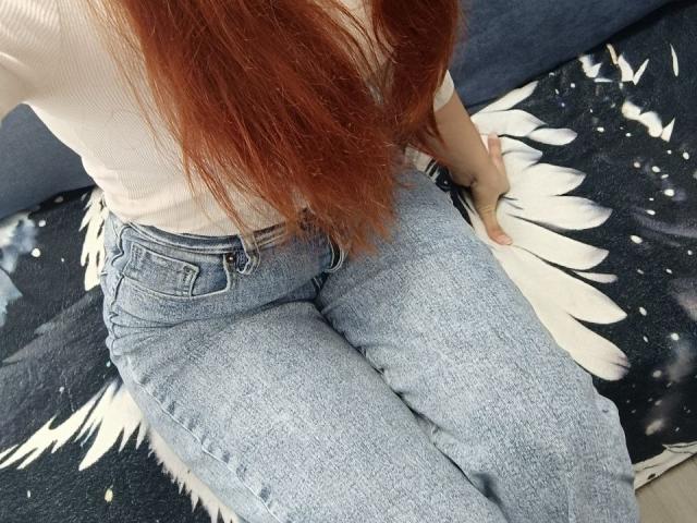 Gvineriyas Hintern in engen, blauen Jeans, weißes Rippstrickshirt, langes, rotes Haar, sitzend auf einem schwarz-weißen Stoff, Hand an der Seite..