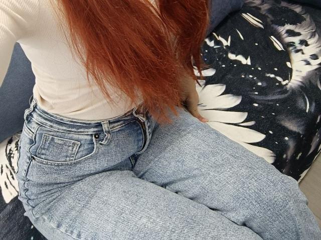 Gvineriya sitzt mit ihrem Hintern in blauen Jeans mit hoher Taille, einem weißen Rippstrickshirt und langen roten Haaren auf einem schwarzen und weißen Sitz mit Blumenmuster..