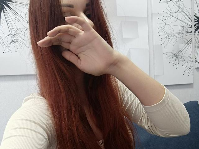Gvineriya with long red hair, covering her mouth with her hand, wearing a beige ribbed top, blue couch in background, white wall with black abstract art.