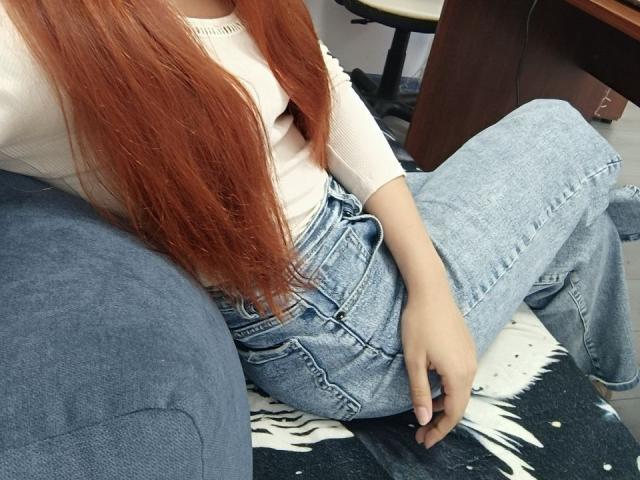 Gvineriya mit langen, roten Haaren, einem weißen Oberteil und hoch taillierten, blauen Jeans, sitzt mit gespreizten Beinen, einer Hand auf dem Oberschenkel, auf einem blau-weißen Sofabezug..