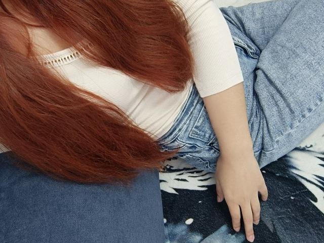 Gvineriya with long, straight red hair sits on a blue and black patterned surface. She wears a white rib-knit shirt and blue jeans. Her right arm is relaxed, fingers slightly spread, while her left arm is partially out of frame. The image focuses on her torso and lower body, with her hair cascading over her shirt. The textures of the rib-knit shirt and denim are clearly visible.