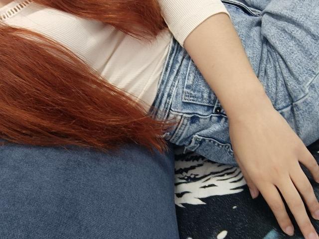 Close-up of Gvineriya's torso and upper legs. Gvineriya has long, straight, red hair that flows over a light pink, ribbed, short-sleeve shirt. She wears high-waisted, blue denim jeans with a slightly wrinkled texture. Her right hand, with a light skin tone and neatly manicured nails, rests on her jeans. The lower part of her jeans shows a black and white patterned fabric beneath. The image focuses on the contrast between the denim and her shirt, and the texture of her hair against the fabric.