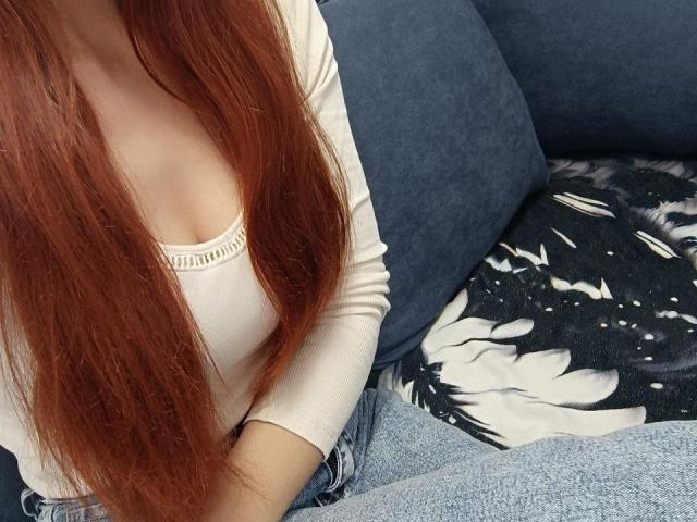 Gvineriya mit langen roten Haaren, einem weißen, gerippten Oberteil, das kleine Brüste zeigt, sitzt auf einem blauen Sofa, trägt blaue Jeans, im Hintergrund ein schwarzes, mit Blumenmuster versehenes Kissen, eine weiße und schwarze, mit Blumenmuster versehene Decke..