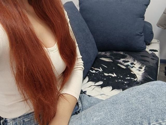 Gvineriya mit langen roten Haaren, einem weißen, gerippten Oberteil und blauen Jeans, sitzt auf einer Couch mit dunkel-weißen, blumigen Kissen. Die Brüste sind teilweise sichtbar..