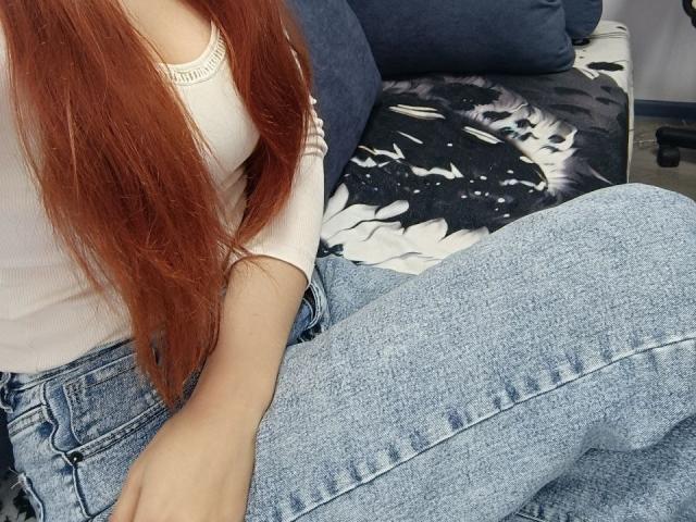 Gvineriya mit langen roten Haaren, einem weißen Oberteil und blauen Jeans, sitzt mit leicht angehobenem Gesäß, wobei ihr Oberschenkel sichtbar ist. Ein schwarz-weißes Kissen ist im Hintergrund zu sehen..