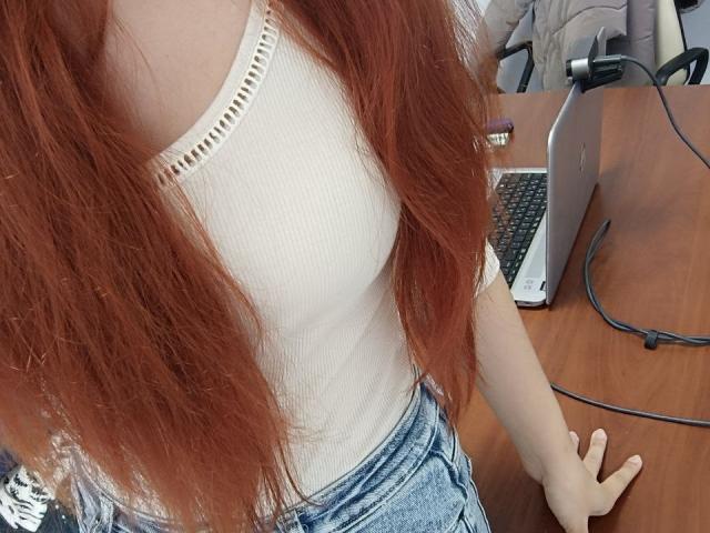 Gvineriya with long red hair, white ribbed tank top, and blue jeans, sitting at a wooden desk with a laptop and black cable. Boobs slightly visible under top.