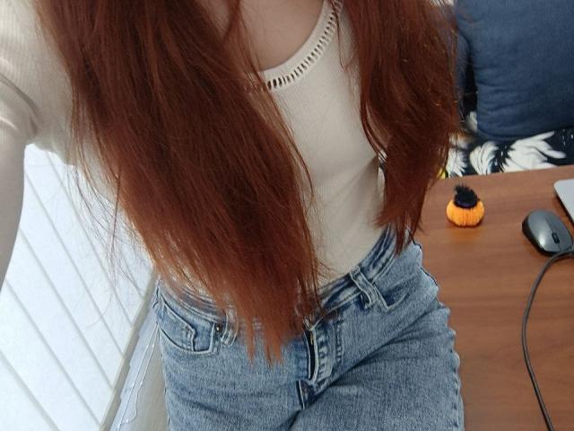 Gvineriya mit langen roten Haaren, einem weißen, gerippten Oberteil und blauen Jeans mit hoher Taille, sitzt auf einem Holztisch mit einer schwarzen Maus und einem orangefarbenen, runden Gegenstand..