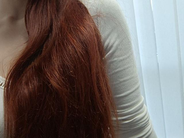 Gvineriyas langes, rotes und leicht gewelltes Haar bedeckt ihre Brüste, die unter einem engen, gerippten, grauen Langarmshirt sichtbar sind. Das Bild konzentriert sich auf das Haar und die Brust..