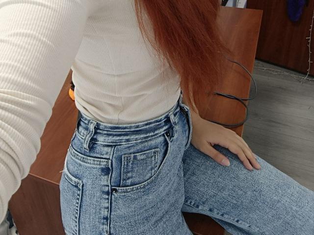 Gvineriya mit roten Haaren, einem weißen, gerippten Hemd und hoch taillierten blauen Jeans, mit der Hand auf dem Oberschenkel, sitzend auf einer braunen Holzfläche. Sichtbarer Hintern, Jeansstoff, lange Ärmel..