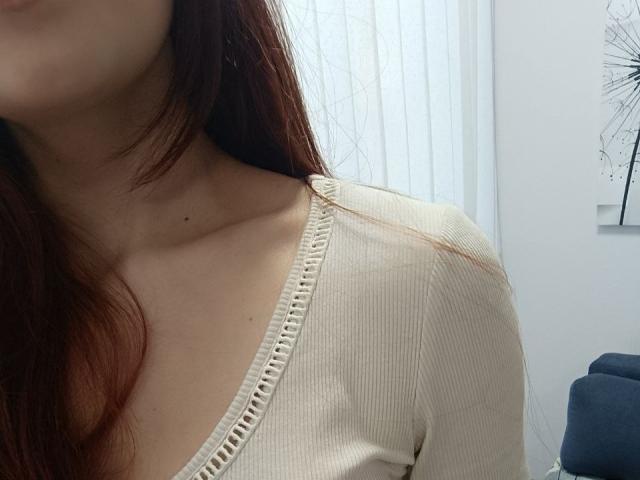 Close-up of Gvineriya's upper body, showing her left shoulder and collarbone. She has fair skin and long, straight, brown hair. Gvineriya is wearing a cream-colored, ribbed, V-neck top with a decorative, woven edge. The top's texture is slightly rough, and a small, dark mole is visible on her right collarbone. The background is blurred, with white vertical blinds and a piece of minimalist art on the right. The image has a casual, everyday feel.
