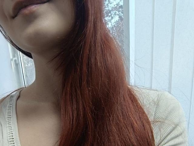 Gvineriya's face with long red hair covering one side, pale skin, and a slightly parted mouth. She's wearing a beige, ribbed, V-neck top. Her tits are partially visible.