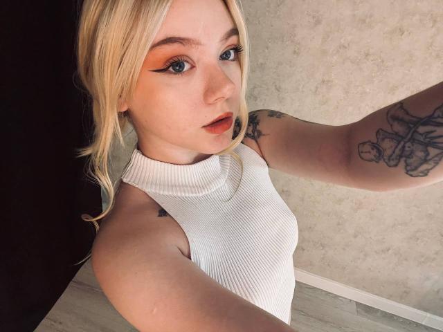 EnchantingEyes with blonde hair, orange eyeliner, and white ribbed top, showing tattoos on her arm, holding a selfie. She has small tits and a cute face.