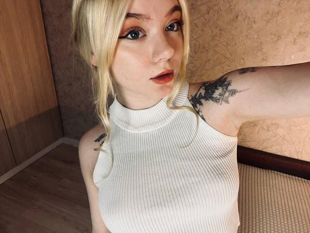 EnchantingEyes with blonde hair, wearing a white ribbed sleeveless top, showing tattoos on her arm, has large blue eyes, and is looking at the camera with slightly parted lips.