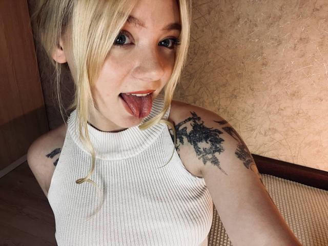 EnchantingEyes, blonde, white ribbed sleeveless top, tattoos on both shoulders, tongue out, blue eyes, fair skin, wooden door in background, selfie, upper body, slight smile, messy hair.