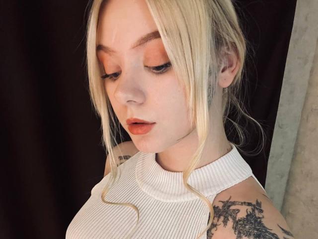 EnchantingEyes with blonde hair, white sleeveless turtleneck, orange eyeshadow, and tattoos on her arm, looking down with slightly parted orange lips.