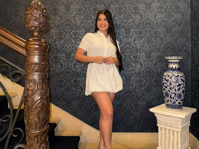 CamilaSofiia with long brown hair, white short-sleeve dress, standing, smiling, holding hands, blue-patterned wall, wooden staircase, ornate pillar, blue-and-white vase, beige floor, wearing pink heels.