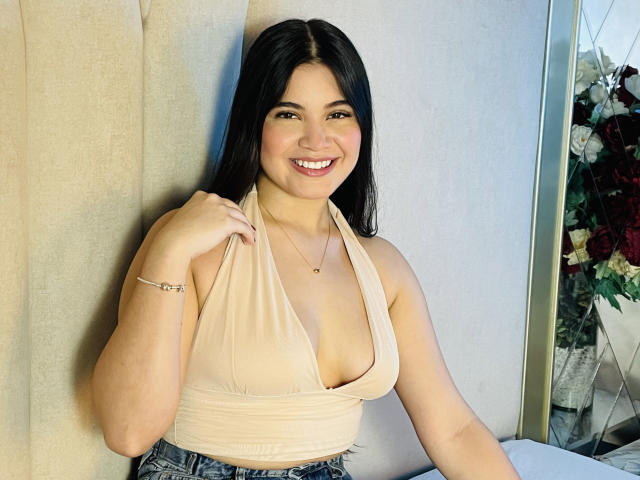 Photograph of CamilaSofiia with long black hair, smiling widely. She's wearing a beige halter-top with a deep V-neck, revealing cleavage. She has a fair skin tone, light makeup, and is sitting against a beige curtain. Her right hand pulls the top slightly. She's wearing a silver bracelet and a small gold necklace. The background has a reflective surface with floral arrangements on the right. She has a full-figured build. The image is brightly lit, emphasizing her cheerful expression.