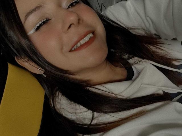 O rosto da Candy, com um sorriso, olhos castanhos, delineador branco, cabelo preto liso, moletom branco, assento amarelo e brincos fofos, mostrando um pequeno conjunto de dentes brancos..