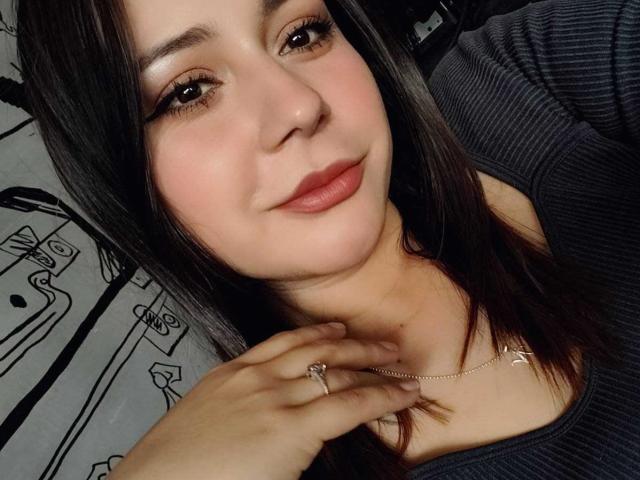 CandyI heeft een lichte huid, donkerbruin haar en roze lippenstift. Ze draagt een grijze top, een ketting en een ring. Haar hand raakt haar nek. Ze glimlacht zachtjes..
