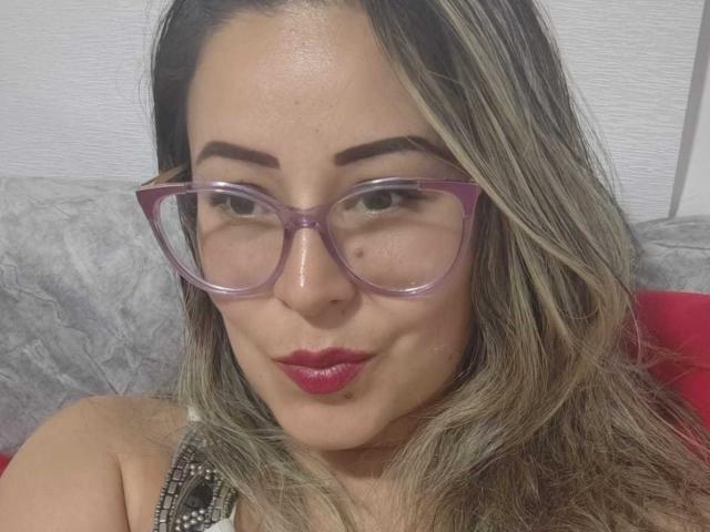Contelaya with purple glasses, red lipstick, and wavy blonde hair, pouting slightly, has a cute face and medium tits. She's sitting on a gray, textured sofa.