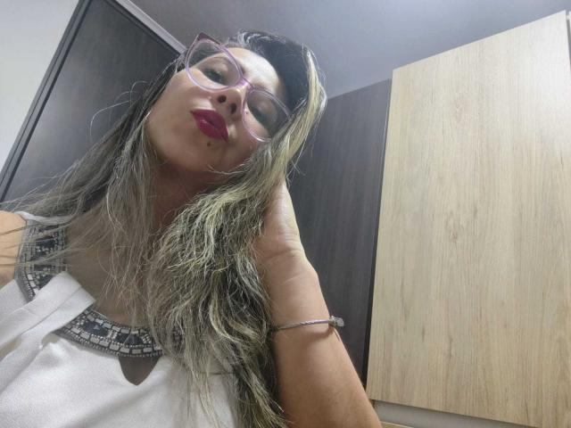 Contelaya with long, wet, blonde-green hair, wearing glasses and red lipstick, looking down at the camera, showing her white top with black bead trim, and a bracelet.