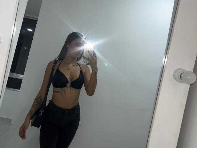 DamiAmbani with long black hair, wearing a black bra and black pants, taking a mirror selfie. Flash from phone covers her tits. Tattoo on right arm.