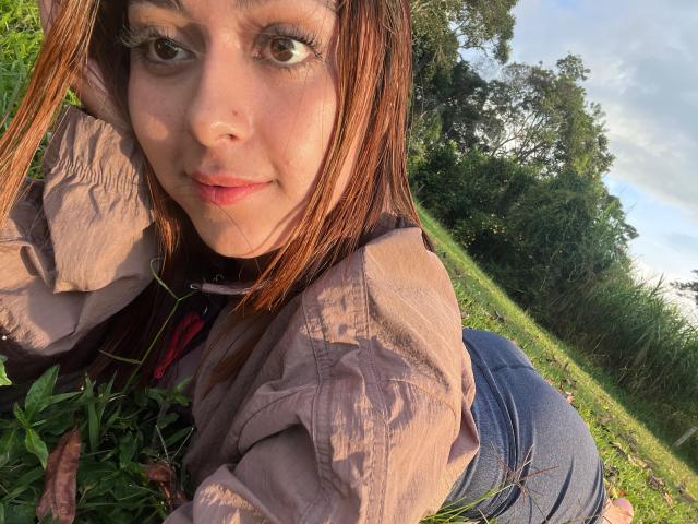 DamiAmbani with brown eyes, pink shirt, and blue jeans, crouching in grass, showing her ass, looking at camera with a slight smile, sunlight on her face.